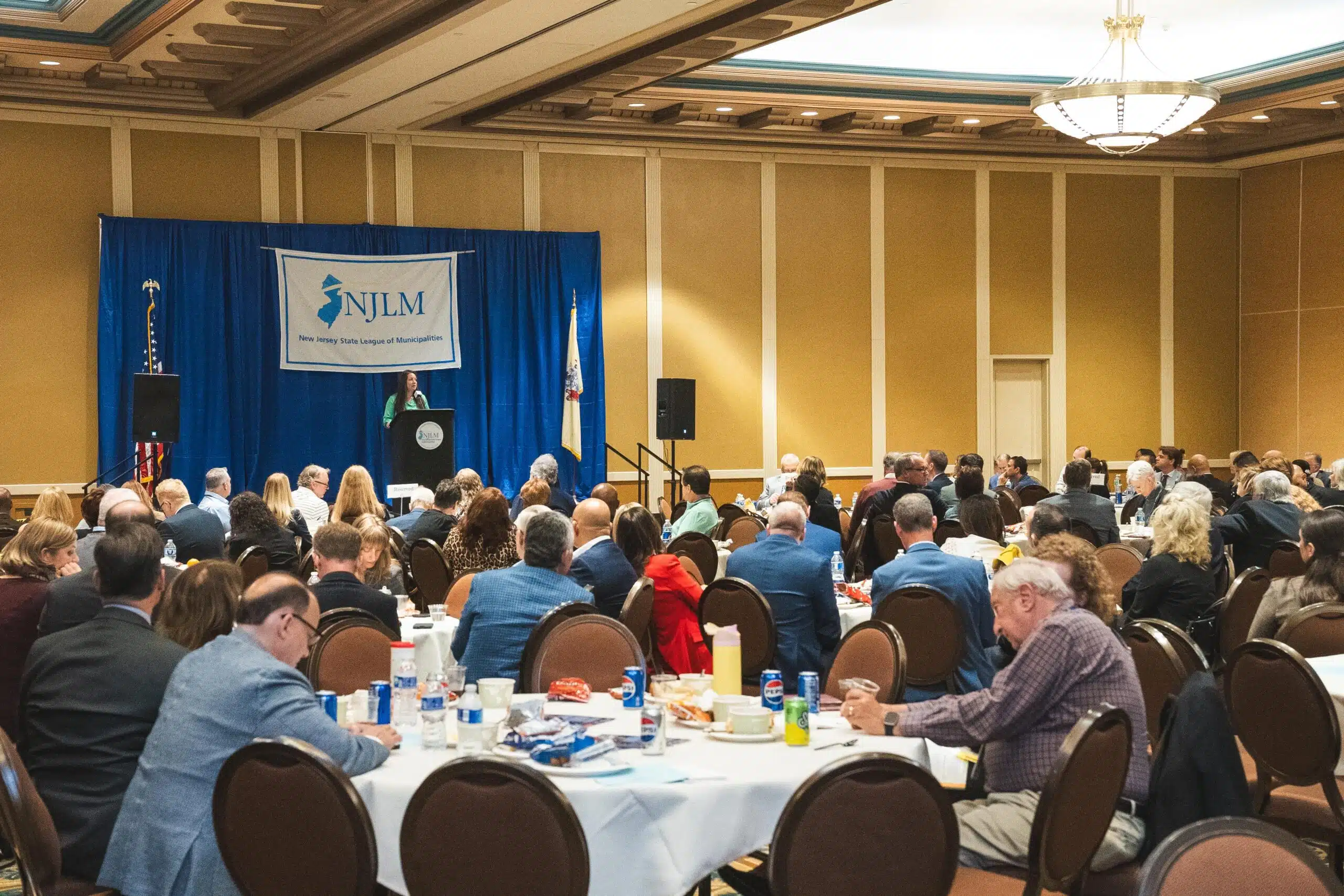 Large room with a lot of people seated at roundtables and a stage with speaker is in the background.
